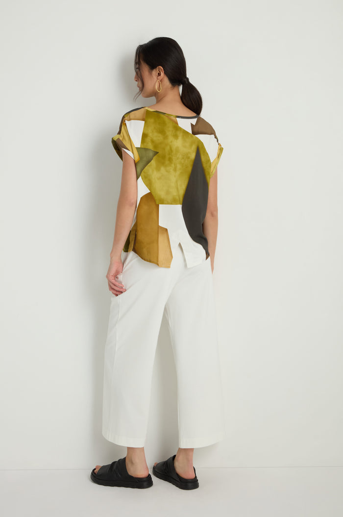 A woman stands against a white wall, facing away, wearing the Collage Print Top in a relaxed fit with geometric patterns, paired with white wide-leg pants, black sandals, and large hoop earrings.