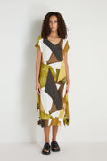 A woman stands against a plain white wall wearing a loose, knee-length Collage Print V-Neck Dress featuring an abstract geometric pattern in olive, black, white, and brown, paired with black sandals.
