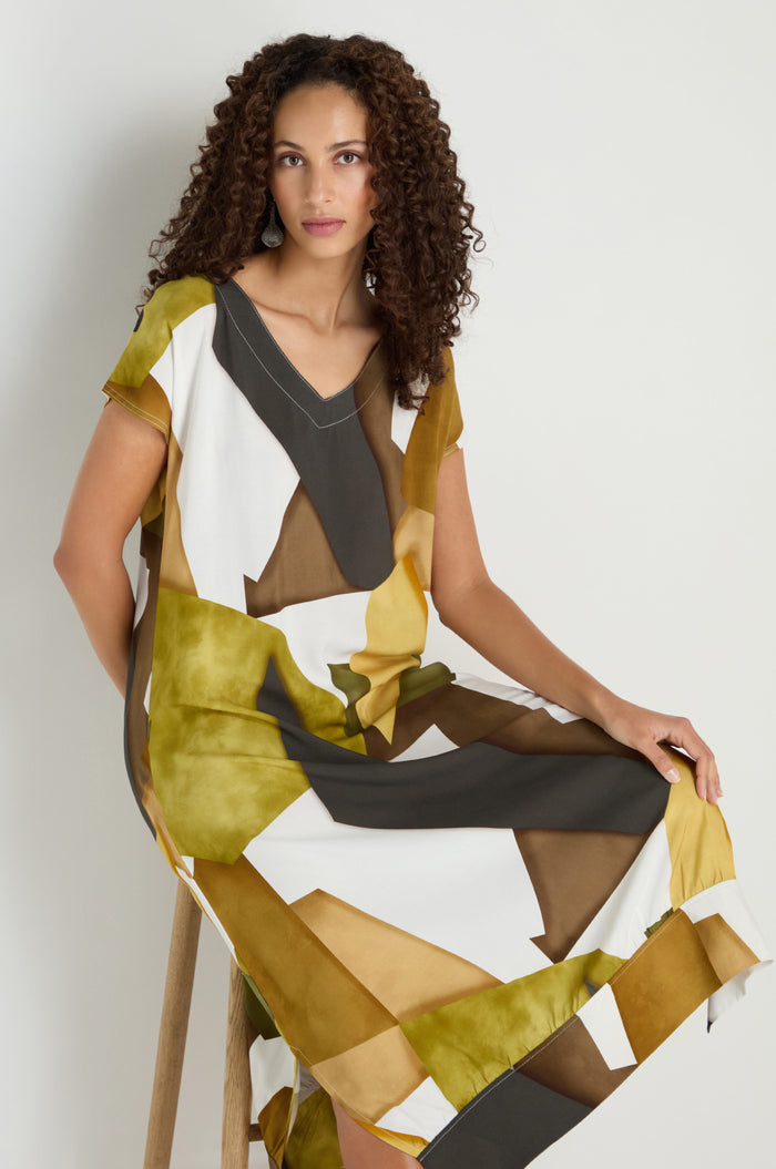 A woman with curly hair sits on a wooden stool against a neutral background, wearing a loose-fitting, short-sleeved Collage Print V-Neck Dress in green, brown, mustard, and white.