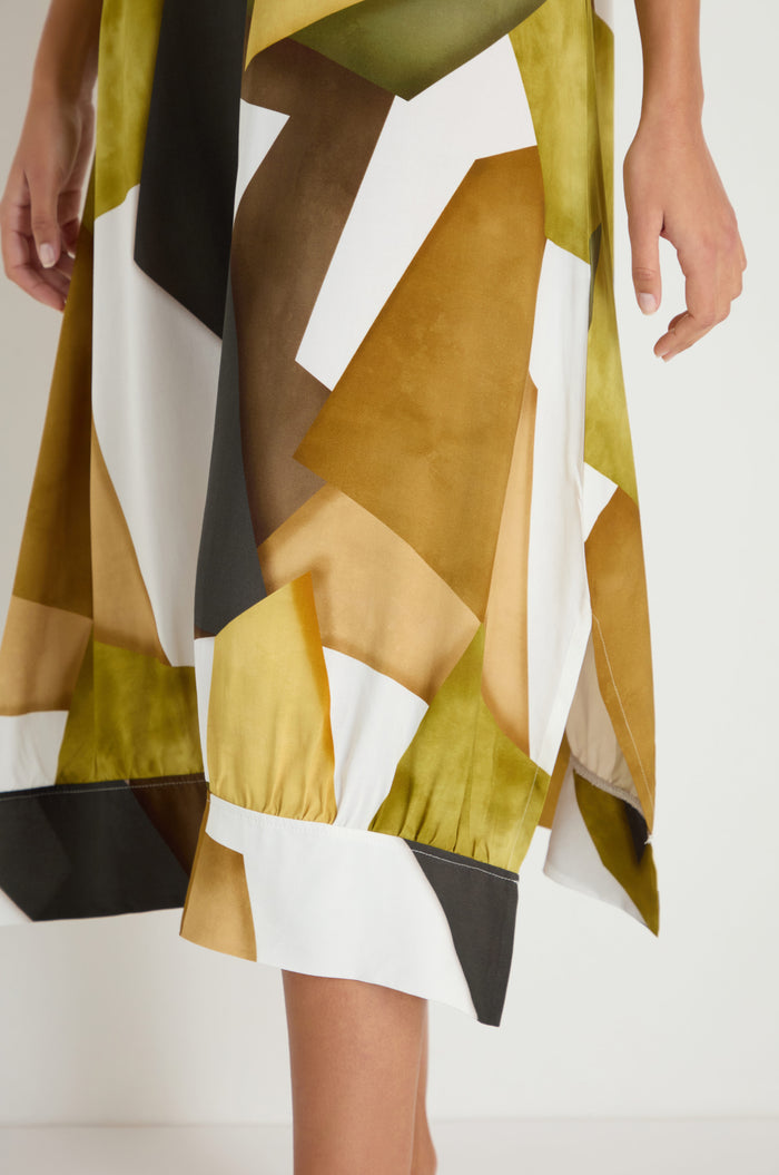 A person wears the Collage Print V-Neck Dress, featuring a geometric pattern in green, brown, white, and yellow. The dress is shown from mid-thigh to calf with one hand visible at the side.