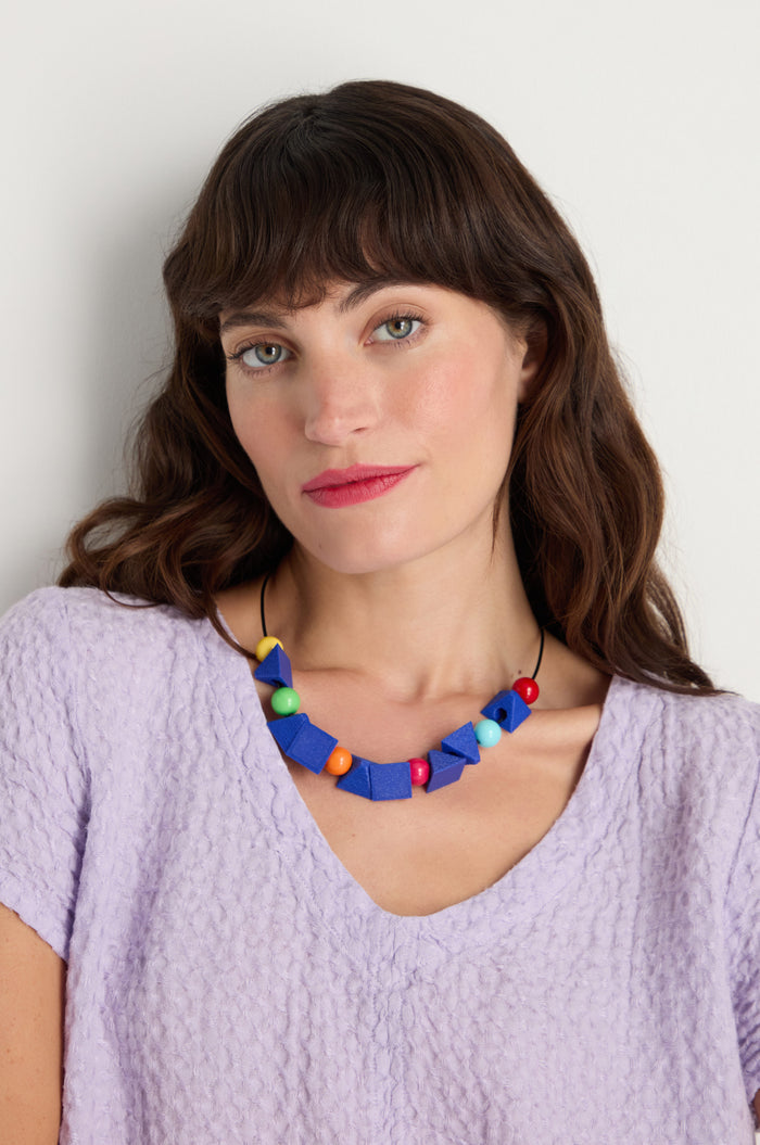 Woman with wavy brown hair wears a textured lavender top and the Colour Pop Shapes Necklace, handmade in Paris, standing against a plain white background.