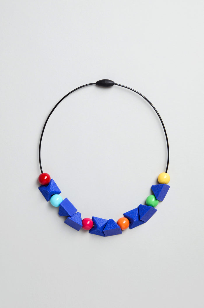 The Colour Pop Shapes Necklace is a handmade geometric statement piece from Paris, with a black wire, large blue beads, and colourful round beads in red, yellow, turquoise, and orange on a plain light background.