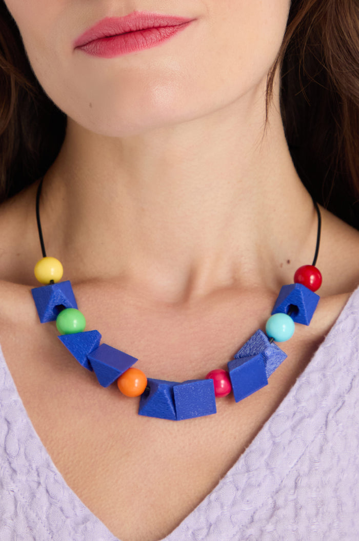 Close-up of a woman wearing the Colour Pop Shapes Necklace, a geometric statement piece with blue and colorful round beads, paired with a light purple textured top. Handmade in Paris, it adds an artistic flair to her outfit.