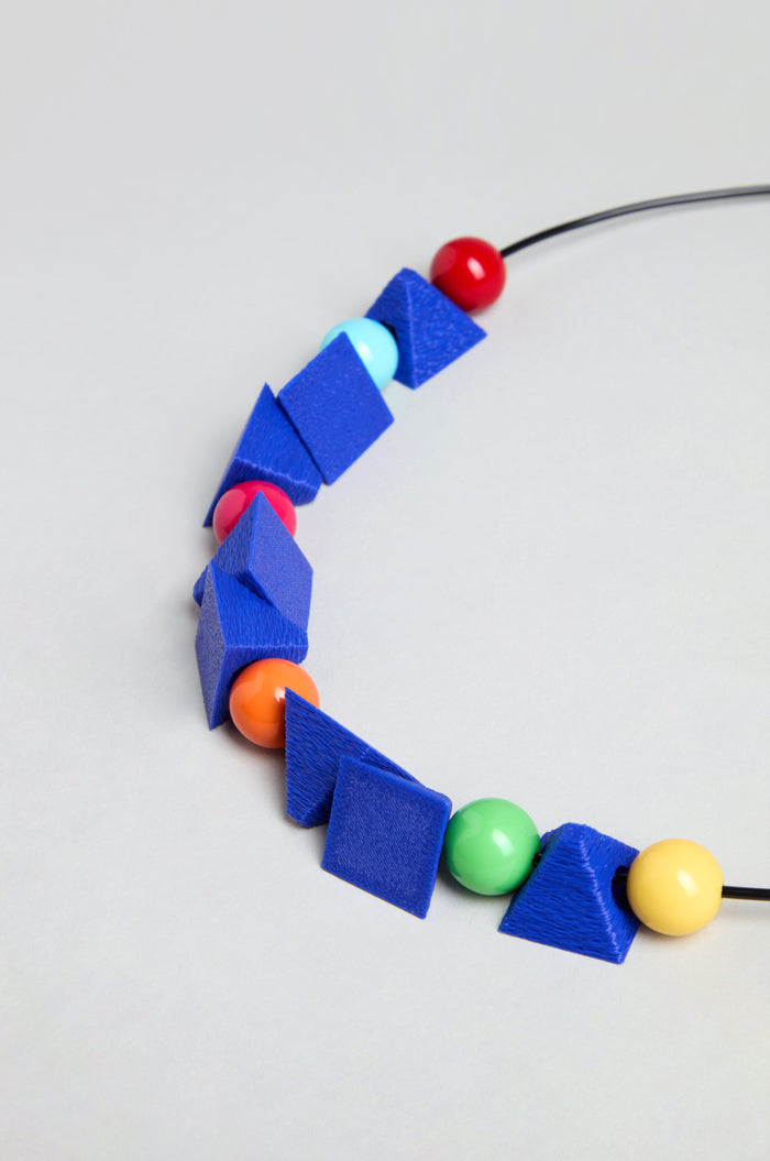 The Colour Pop Shapes Necklace is handmade in Paris, featuring large geometric blue shapes alternating with round red, yellow, green, orange, and blue beads on a black cord, displayed against a plain background.