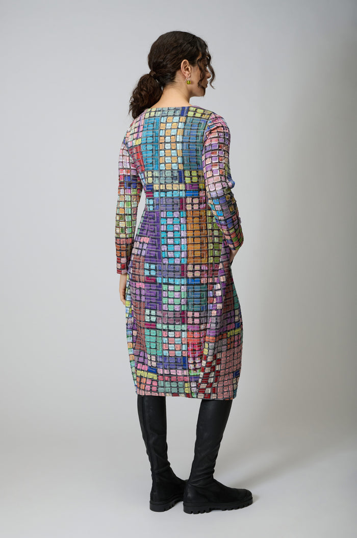 A person with curly hair in a ponytail wears the Coloured Glass Tiles Jersey Bubble Dress with long sleeves and a checkered pattern, paired with black knee-high boots, facing away from the camera against a plain background.