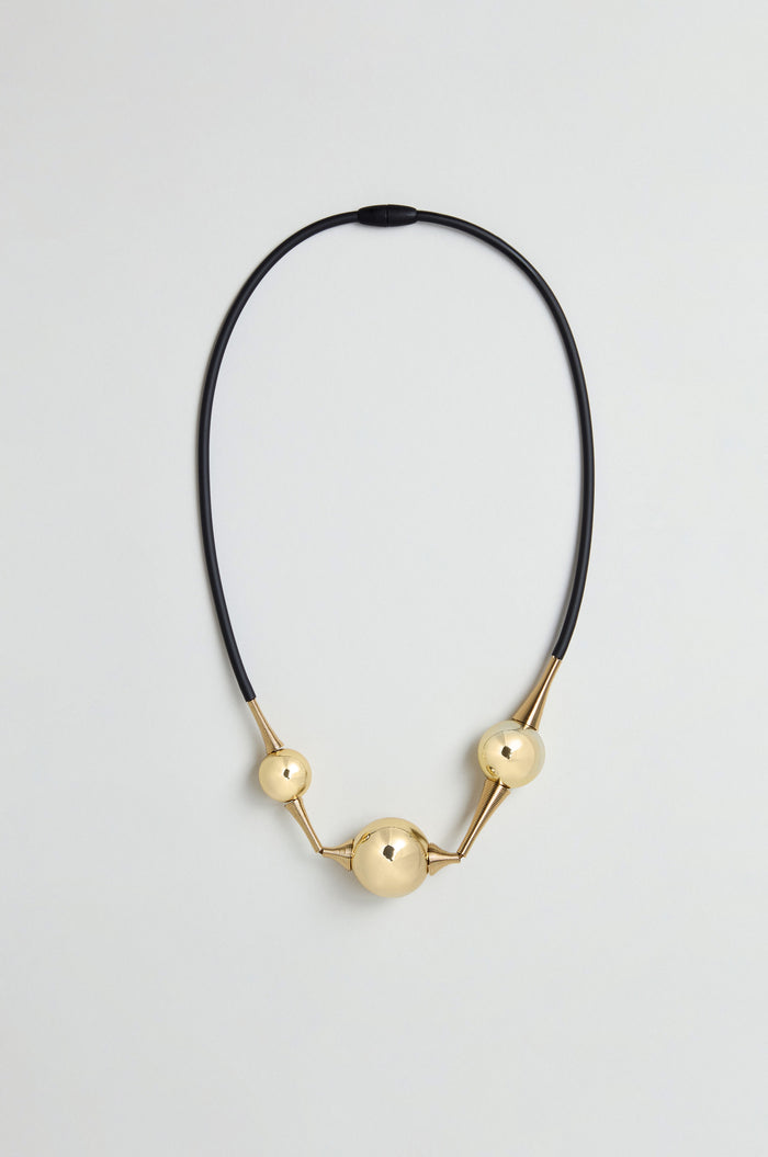 The Cosmos Necklace features three gold spherical beads of varying sizes arranged along a black silicone cord, creating a sculptural jewellery piece displayed against a plain light background.