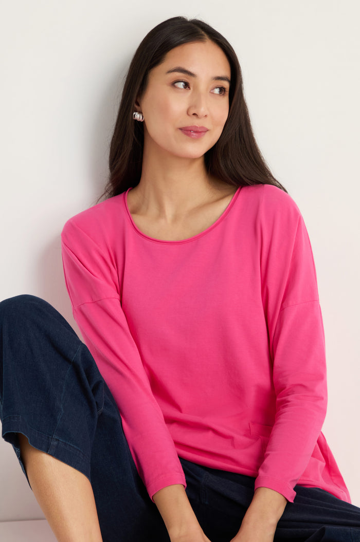 Woman with long dark hair wearing the Cotton Jersey Boxy Pocket Top in bright pink and dark pants sits against a light wall, looking to the side with a slight smile.