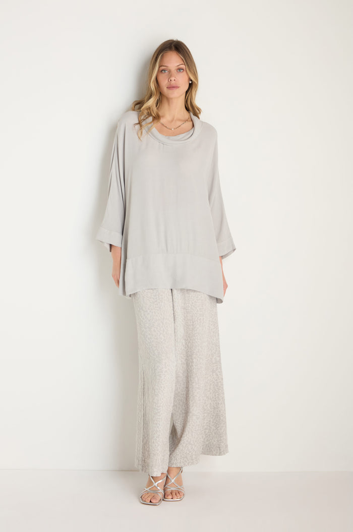 A woman stands against a white wall wearing a loose, light gray Crepe Boxy Top, wide-leg patterned light gray MTT5245-LVC pants, and strappy sandals.