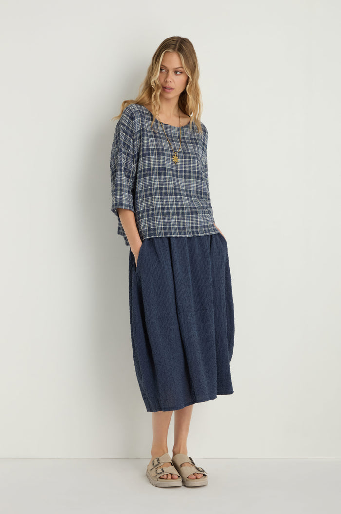 A woman stands against a white wall wearing a blue plaid top, beige sandals, and the Crinkle Bubble Skirt—a dark blue skirt with an elasticated waist—her hands in her pockets.