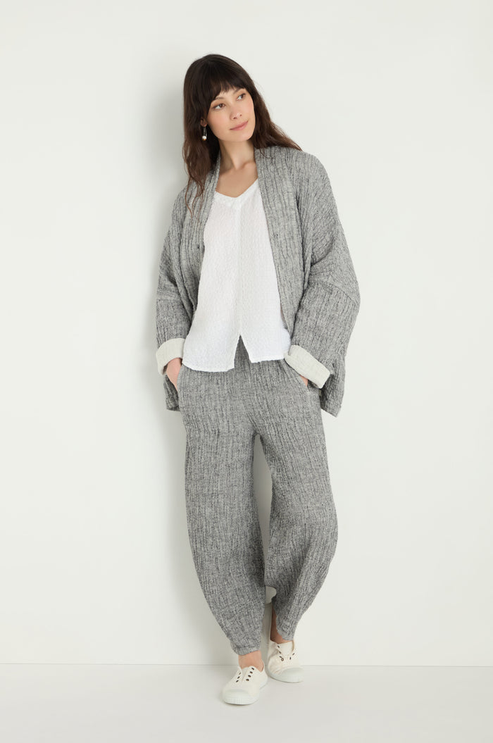 A woman stands against a white wall wearing a loose grey knit cardigan, Crinkle Double Face Linen Bubble Trouser, a white blouse, and white sneakers.
