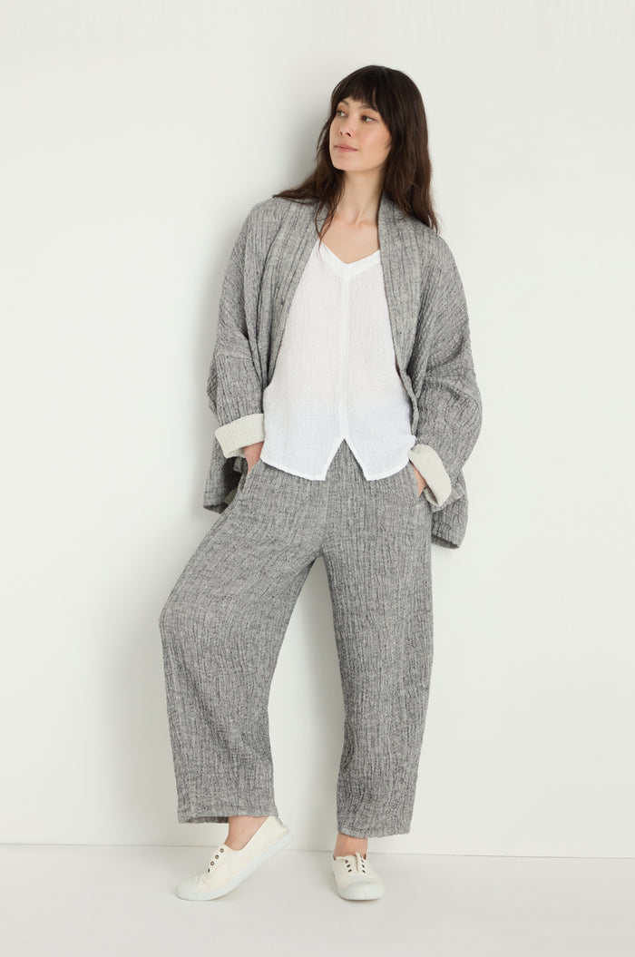 A person stands against a white wall wearing Crinkle Double Face Linen Bubble Trousers in grey, styled loosely over a white shirt and paired with white sneakers.