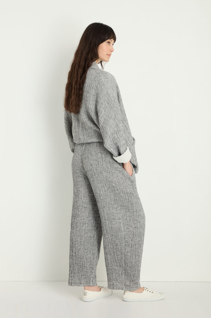 Person stands against a white wall, wearing Crinkle Double Face Linen Bubble Trouser in textured grey with white sneakers, hands in pockets and facing slightly away from the camera.