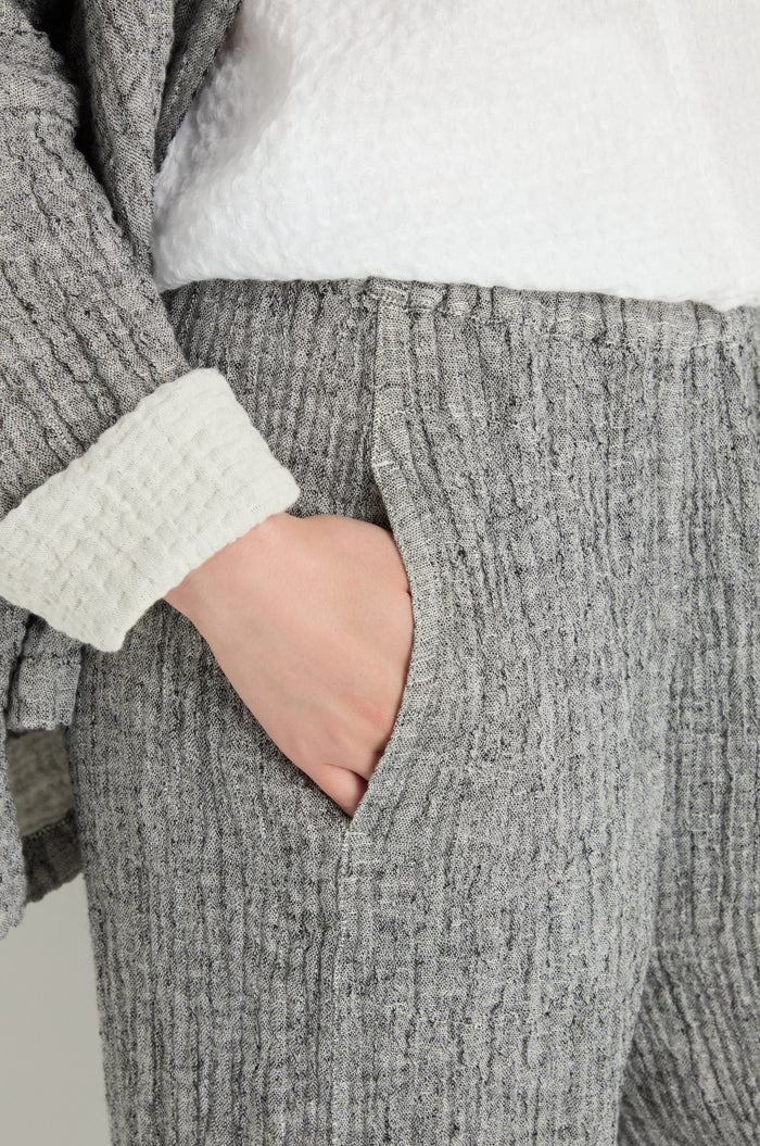 A person wears the Crinkle Double Face Linen Bubble Trouser in textured grey linen with a white top and has their hand in the pocket.