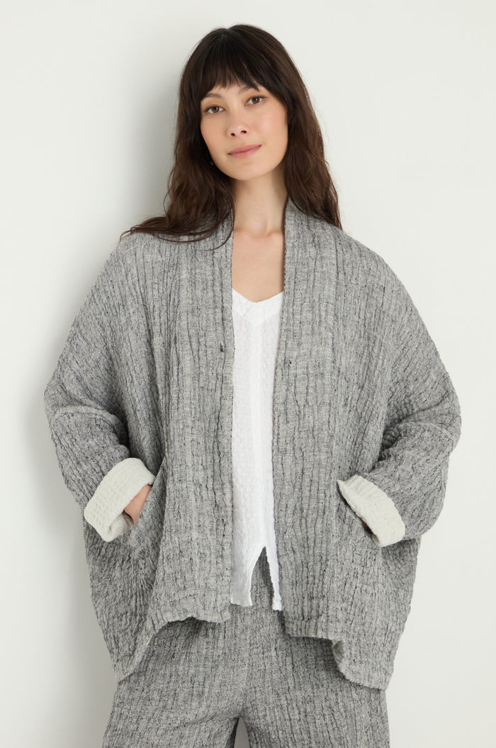 A woman with long brown hair wears the Crinkle Double Face Linen Jacket in gray over a white top and matching pants, standing against a plain light background.
