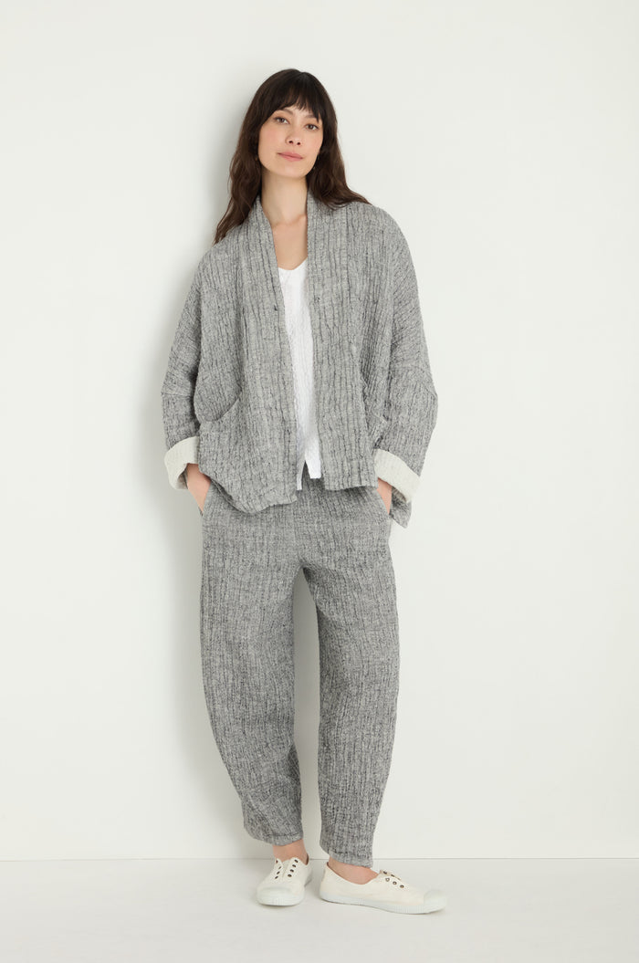 A person stands against a white wall wearing the Crinkle Double Face Linen Jacket in gray, paired with matching loose pants, a white shirt, and white sneakers, hands in pockets.