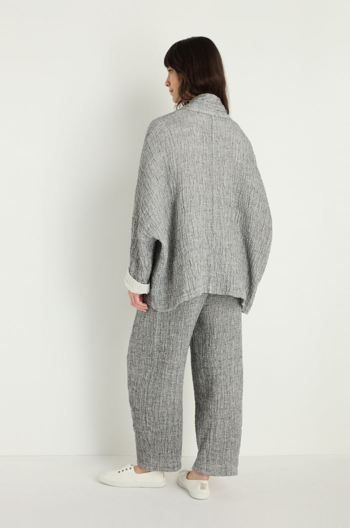 A person faces away from the camera against a plain background, wearing the Crinkle Double Face Linen Jacket in gray with matching pants and white casual shoes. The outfit is loose-fitting with a distinctive crinkle texture.