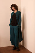 Person wearing a teal textured fabric long jacket and matching Crinkle Linen Blend Bubble Trousers with an elasticated waist over a black shirt, standing indoors against a beige wall, hands in pockets and maintaining a neutral expression.