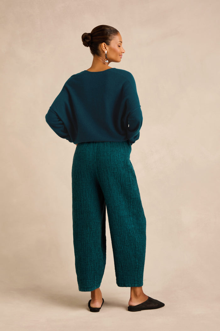 A woman stands with her back to the camera, wearing a teal sweater and the Crinkle Linen Blend Bubble Trousers in textured teal, paired with black flats. Her hair is tied back, and her hands are in her pockets as she poses against a neutral background.