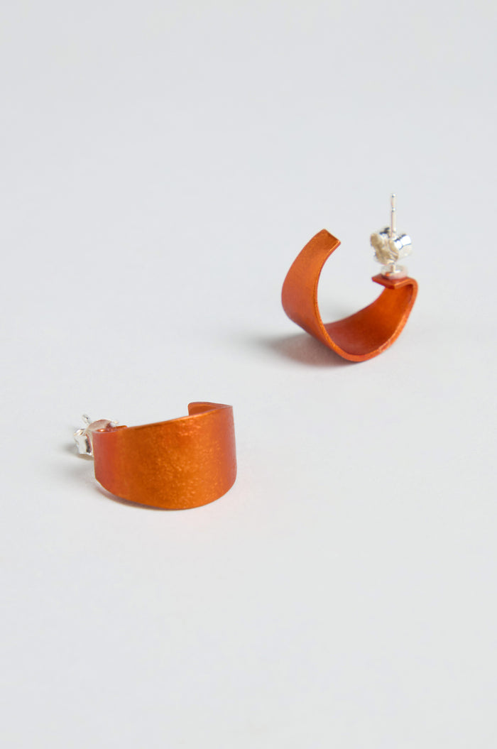 The Curve Earrings feature orange, semi-circular curves made from anodised aluminium with sterling silver posts, shown on a plain white background.