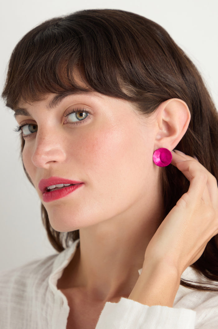 A woman with light skin and brown hair wears a white top and the Curved Disc Earrings—bright pink with sterling silver posts—holding one earring in her hand while looking slightly to the side.