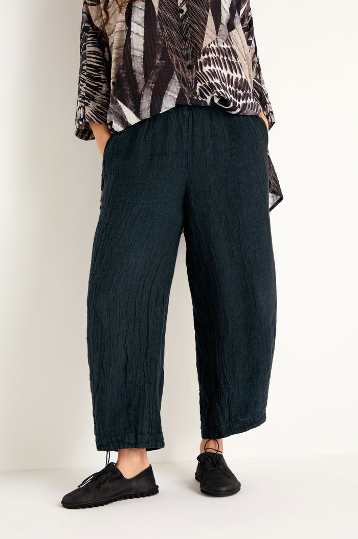 A person wearing the Textured Linen Turquoise Bubble Trousers—crafted from breathable natural linen with a crinkled, wide-leg silhouette—paired with black lace-up shoes and a patterned top, stands hands-in-pockets against a plain white background.