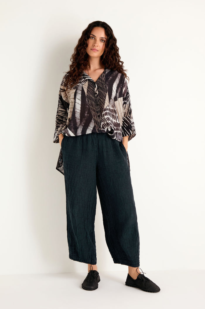 Woman stands against a plain white wall wearing a patterned loose top, Textured Linen Turquoise Bubble Trousers, and black shoes.