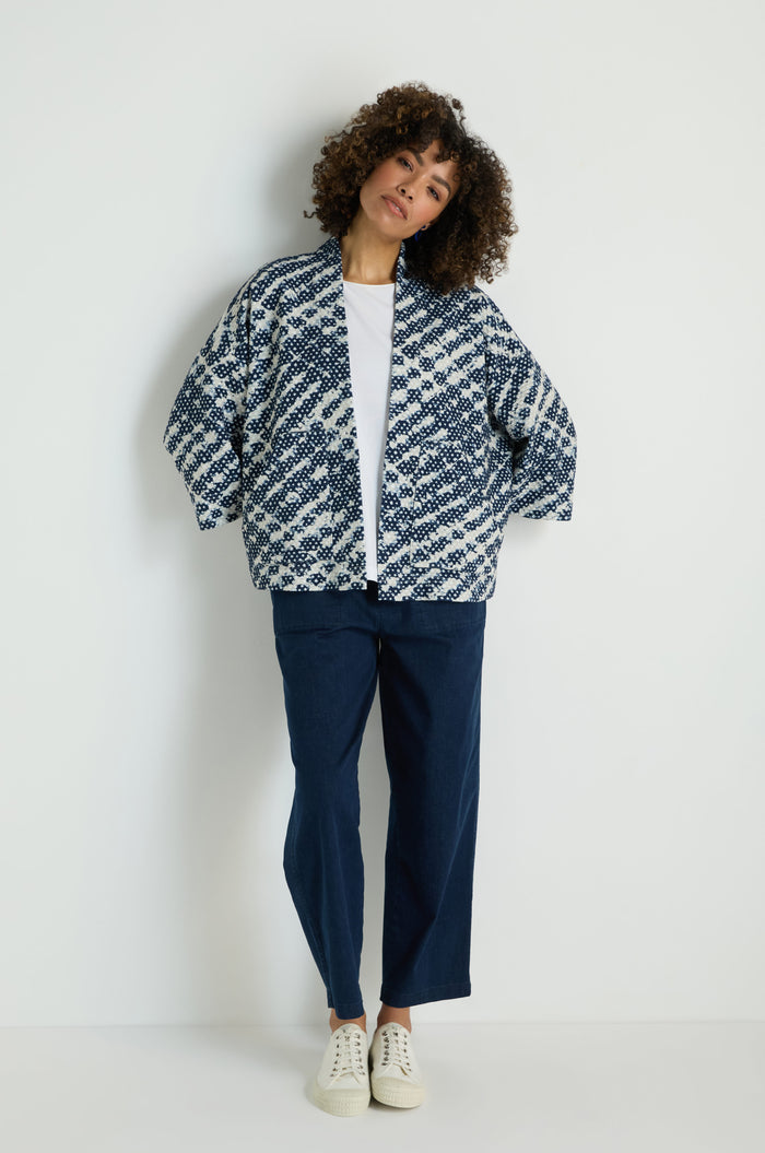 A person stands against a plain white background wearing a white T-shirt, navy blue pants, white sneakers, and the Diamond Tie Dye Jersey Jacket.