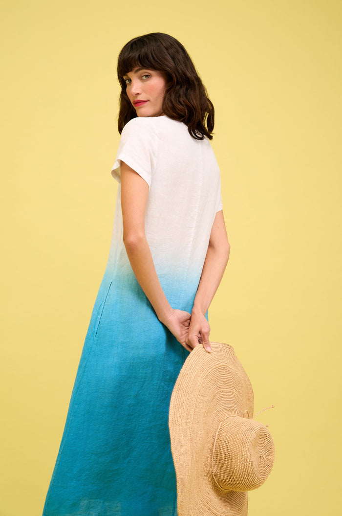 Woman wearing the Dip Dye Linen Dress with short sleeves stands against a yellow background, looking over her shoulder and holding a wide-brimmed straw hat behind her.