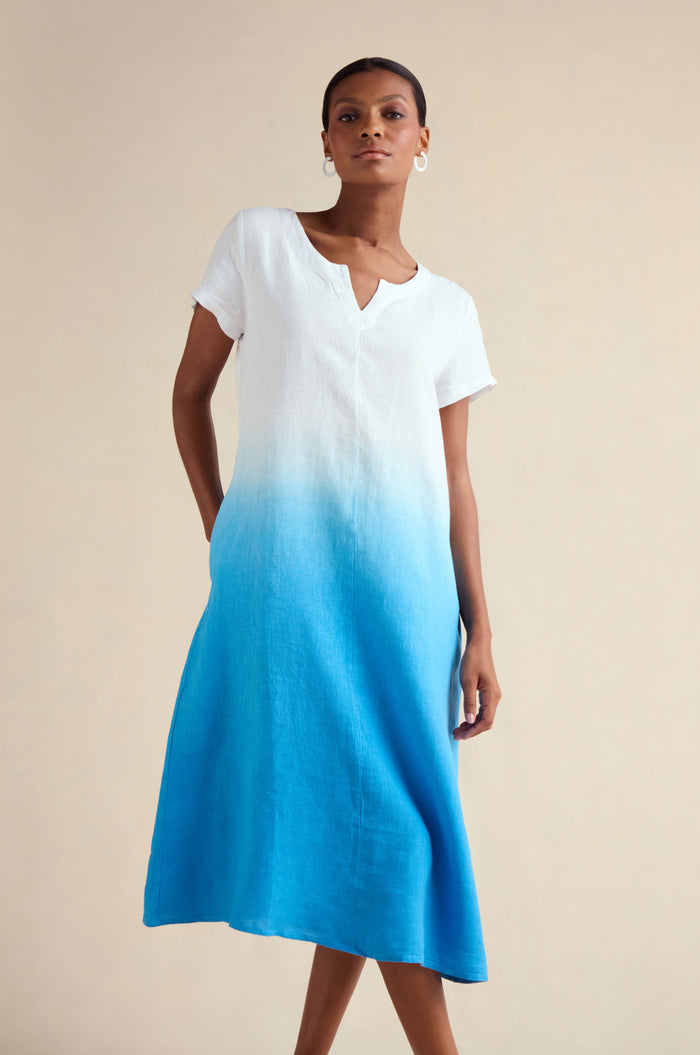 A woman wears the Dip Dye Linen Dress, featuring a short-sleeve V-neck design with a white top blending into a bright blue ombre A-line skirt, standing against a plain light background.