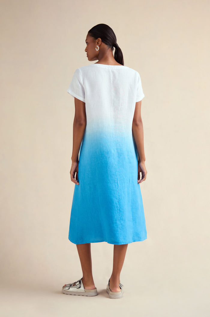 A woman faces away from the camera, wearing the Dip Dye Linen Dress in a white-to-blue gradient A-line style, set against a neutral background with casual shoes.