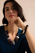 A woman with short dark hair wears a blue top, a chunky dark and metallic necklace, and the striking Dotty Bangle in black wood, resting her chin on her hand as she looks at the camera.