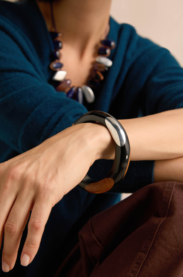 Wearing a dark blue top, brown pants, a chunky blue and white necklace, and the Dotty Bangle—a striking black wood accessory—on their wrist, with one arm resting on their knee.