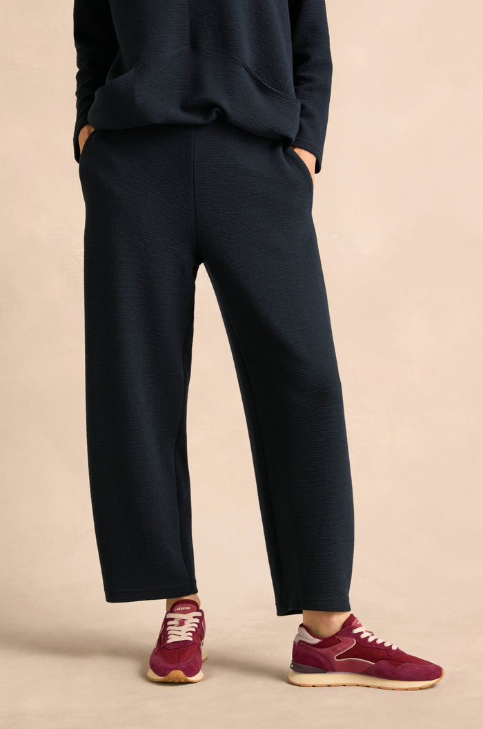 A person is wearing the Double Jersey Crinkle Trousers with a black sweatshirt, hands in pockets, maroon sneakers, and standing on a beige background. These trousers are made in England.