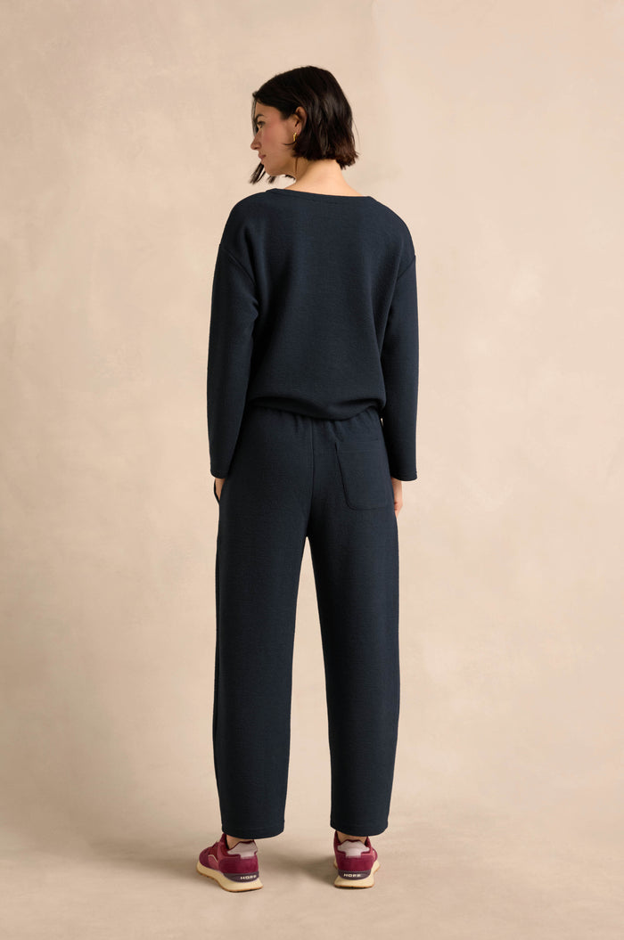 A person stands facing away, dressed in a dark long-sleeve top and the Double Jersey Crinkle Trousers—relaxed-leg trousers crafted in England—paired with maroon sneakers, set against a neutral background.
