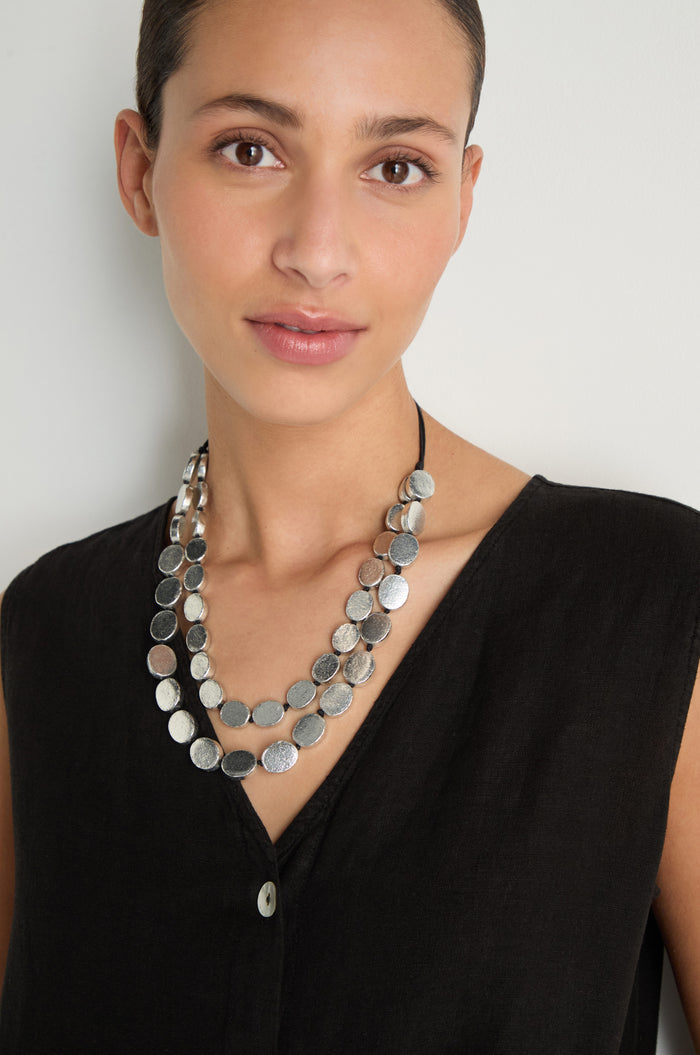 A person with short hair wears a sleeveless black top and the Double Strand Oval Disc Necklace while standing against a plain light background.