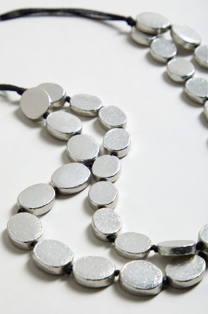 Close-up of two silver metallic beaded necklaces with black cords, including the Double Strand Oval Disc Necklace, arranged on a white surface.