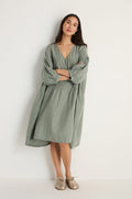 A woman with a neutral expression stands against a white wall, arms crossed, wearing the Easy Fit V-Neck Sage Linen Dress and beige shoes.