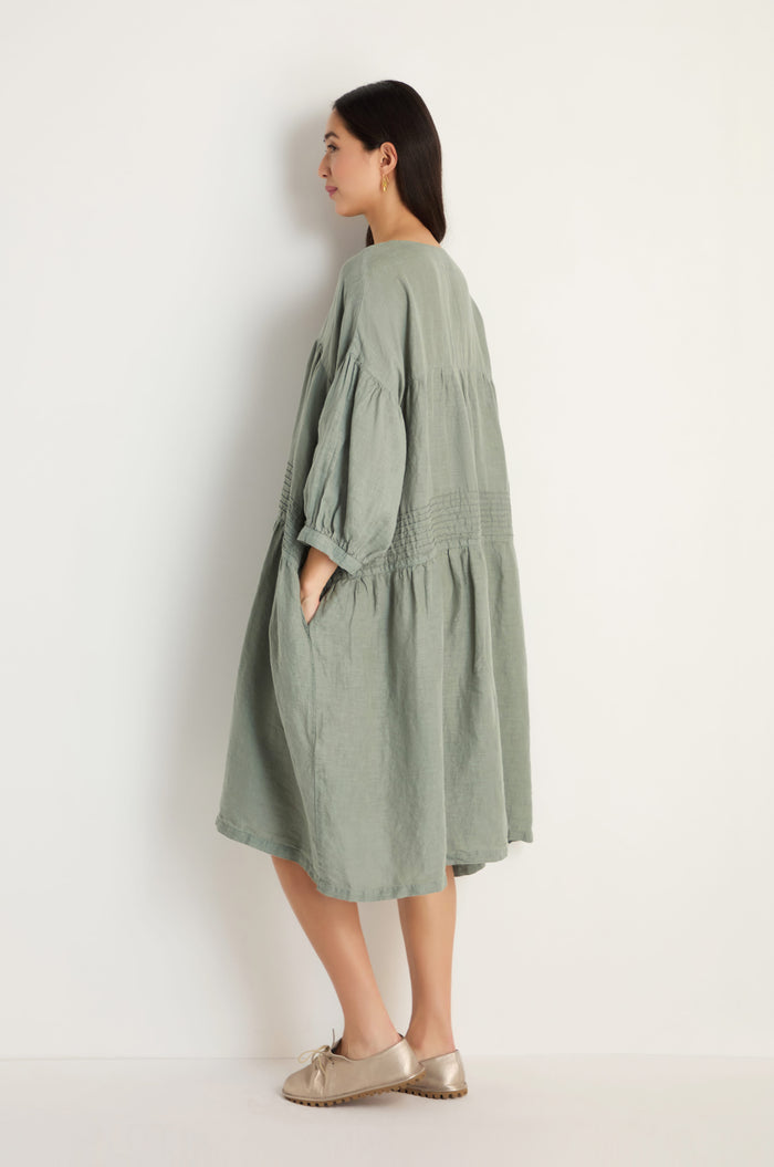 A woman stands sideways against a plain white wall, wearing the Easy Fit V-Neck Sage Linen Dress with pockets and beige shoes.