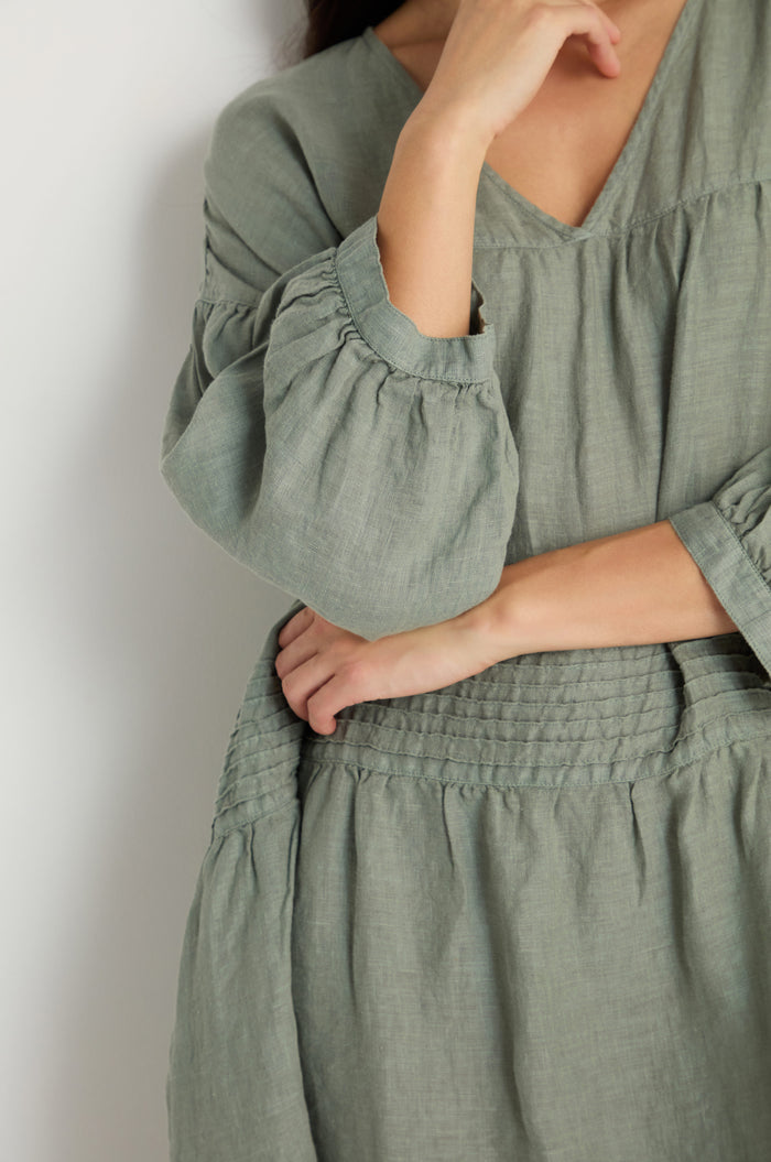 A person is shown from the shoulders down, wearing the Easy Fit V-Neck Sage Linen Dress with puffed sleeves, standing against a plain light background.