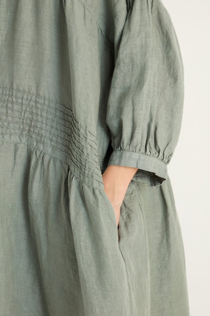 Close-up of a person wearing the Easy Fit V-Neck Sage Linen Dress, showcasing textured stitching, a relaxed A-line silhouette, and one hand tucked into the pocket.