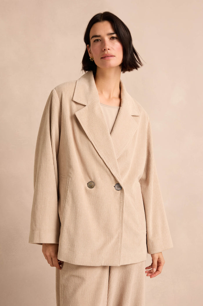 A woman with short dark hair wears the Ecru Jumbo Cord Caban Jacket, featuring pure cotton fabric and two buttons, along with matching pants, as she stands against a light neutral background.