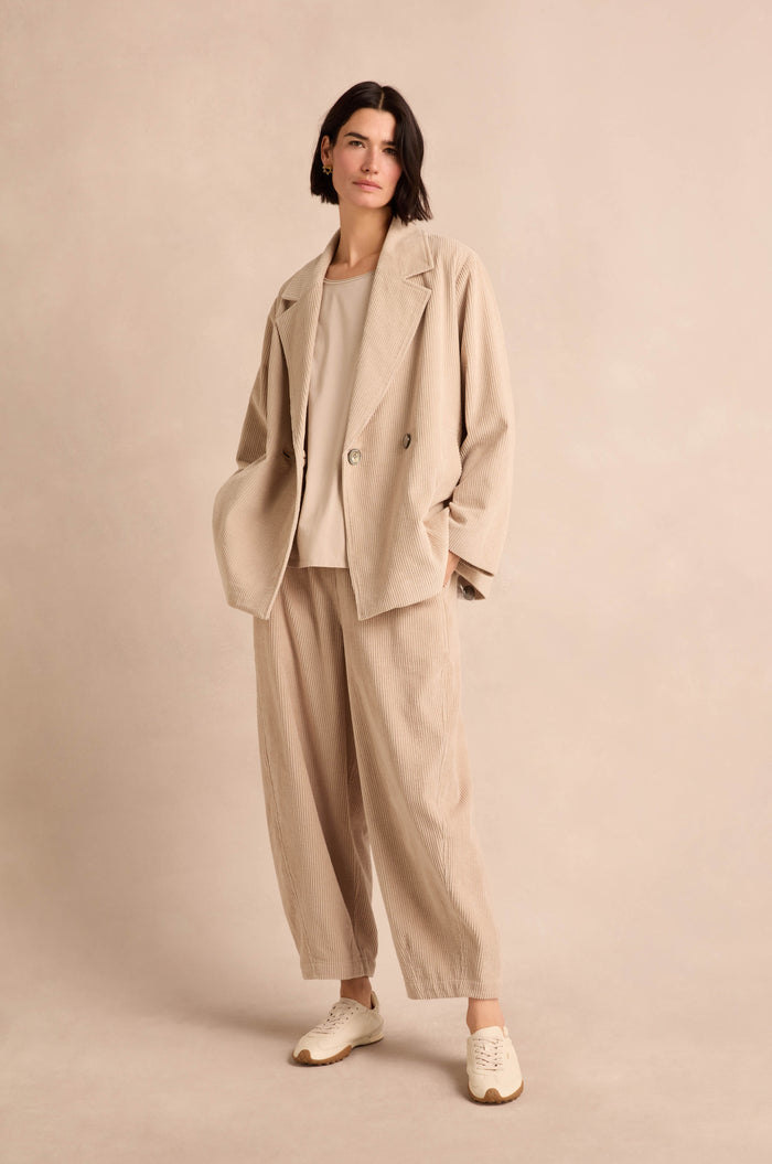 A woman stands wearing the Ecru Jumbo Cord Caban Jacket—a loose, double-breasted style—paired with wide-leg pants, a white top, and cream sneakers against a neutral background.
