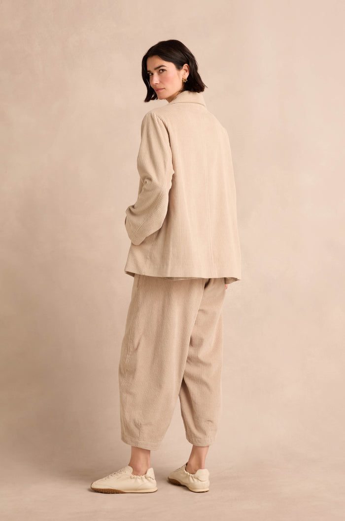 Person standing and looking over their shoulder, wearing an Ecru Jumbo Cord Caban Jacket and white sneakers, against a plain beige background.