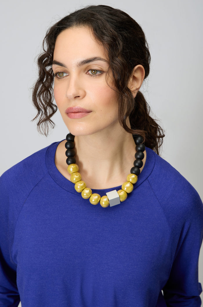 Woman in a blue top wears the Elements Necklace—a chunky, contemporary Christina Brampti piece featuring large yellow and black beads with a silver accent—while gazing slightly to the side against a plain background.