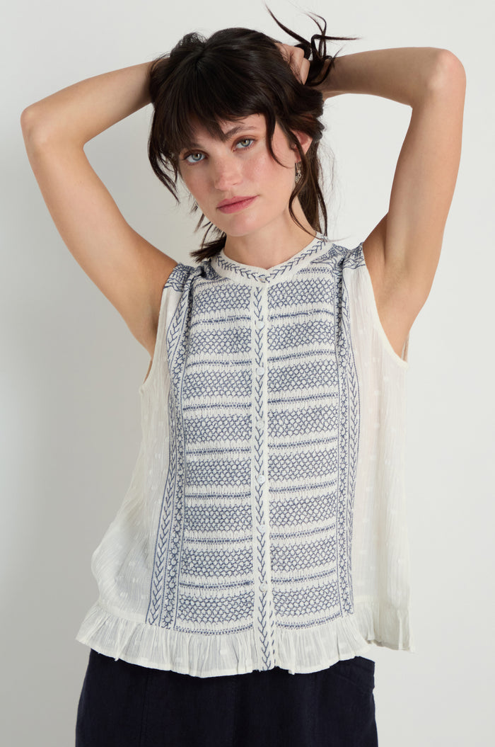 A woman stands with her hands behind her head, wearing the Embroidered Button-Through Top featuring blue patterns and a ruffled hem, set against a plain background.