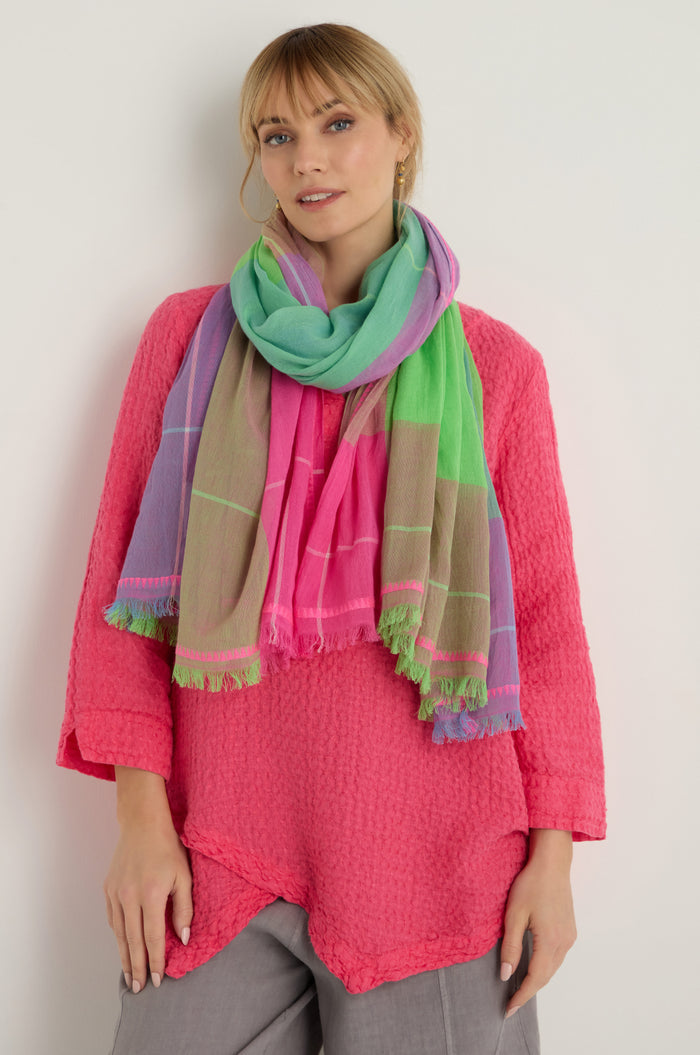 A woman stands against a white background wearing a bright pink textured top, gray pants, and the Embroidered Colour Pop Check Cotton Scarf with green, pink, and purple tones.