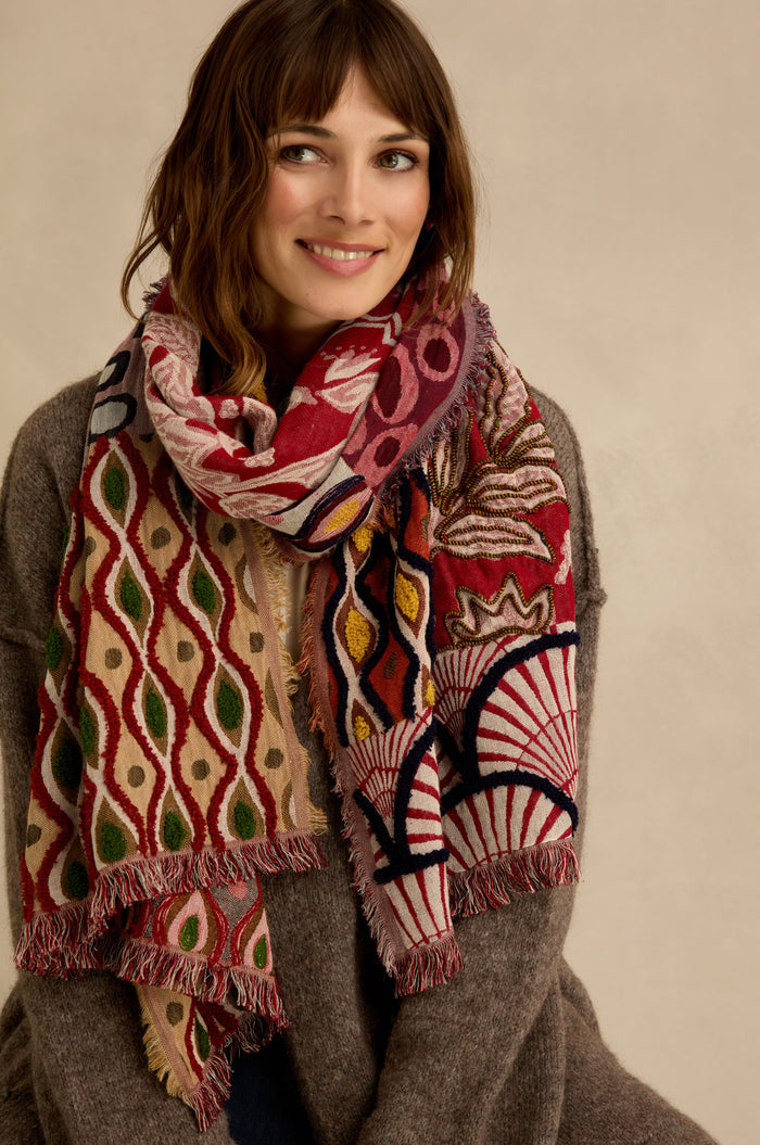 A woman smiles at the camera against a neutral background, wearing a brown sweater and the Embroidered Cotton Wool Scarf, which is adorned with vibrant red, green, and cream geometric and floral motifs. The scarf features delicate fringed edges for an added touch of elegance.