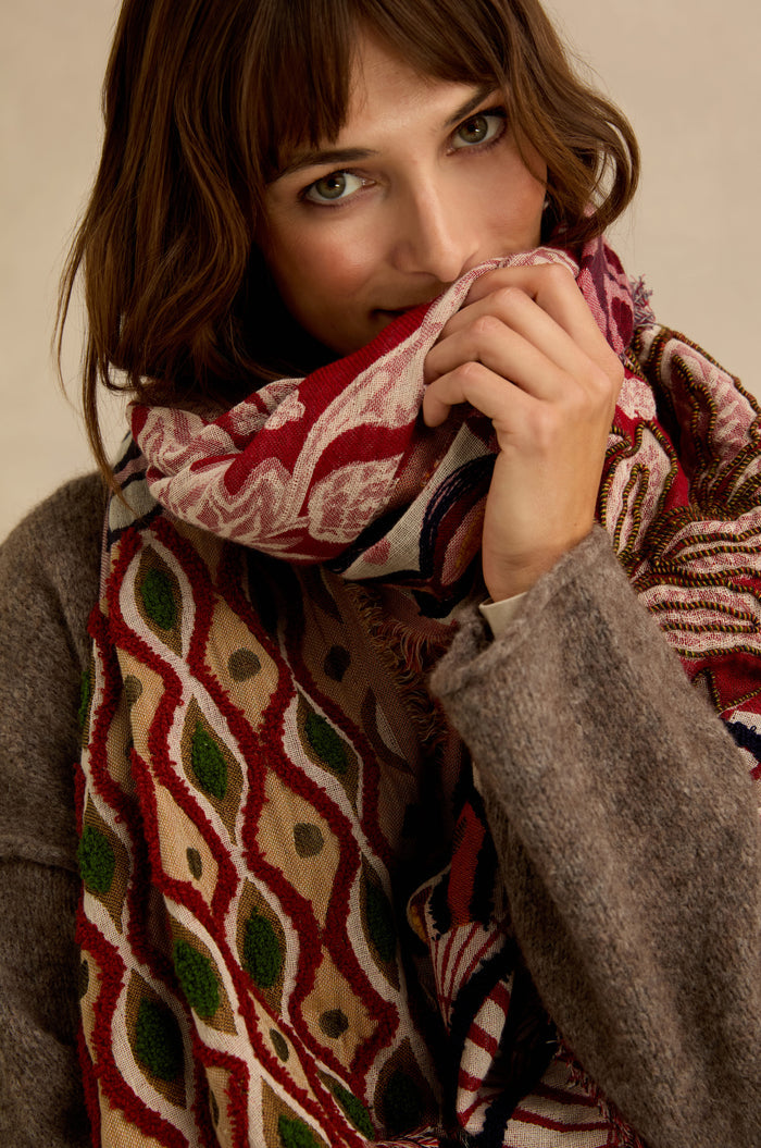 A woman with brown hair in a gray coat looks at the camera, holding the Embroidered Cotton Wool Scarf—featuring large red, green, and beige geometric and floral motifs—close to her face.