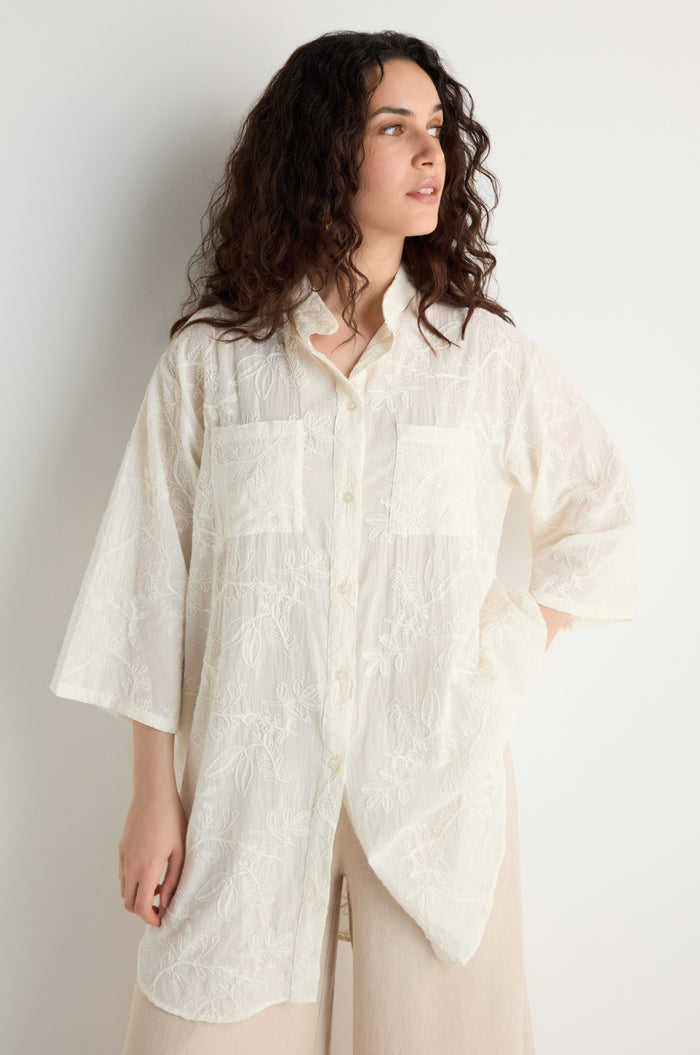 Woman with wavy dark hair wears the Embroidered Long Floral Shirt, a loose, semi-sheer top with floral embroidery and two front pockets, posing against a plain light background.