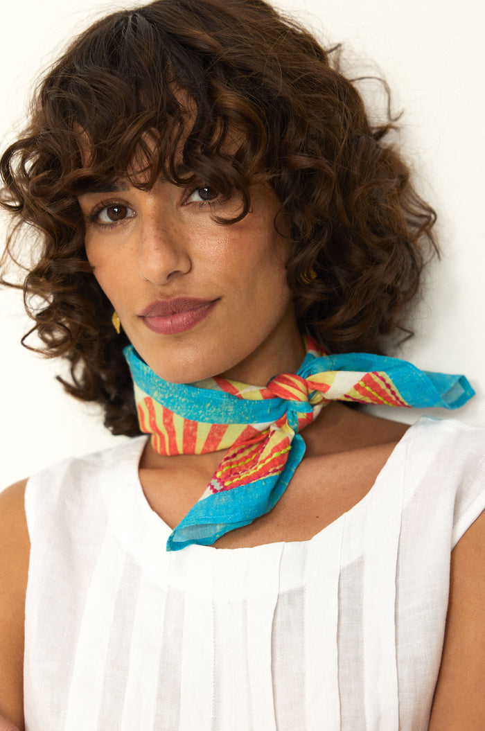 A woman with curly brown hair wears a white sleeveless top and the Embroidered Parasols Cotton Bandana, featuring a colorful blue and red print, tied around her neck as she stands against a plain background.